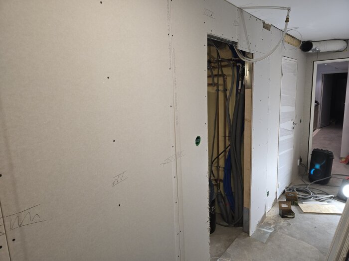En halvfärdig vägg med gipsskivor i en källare, synligt rörsystem, sladdar och ett ljus utplacerat på golvet.