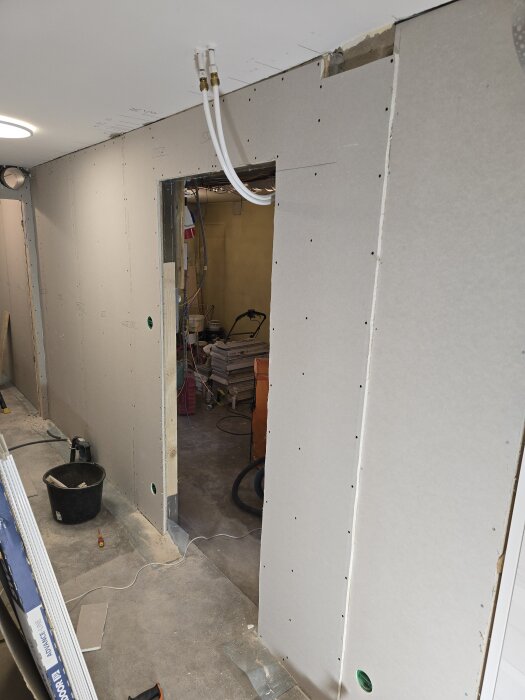 Vägg under konstruktion med gipsplattor och en öppning för dörr, synliga kablar i taket, verktyg och material på golvet.