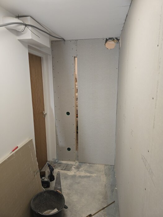 Korridor under renovering med gipsskivor på vägg, ventilation och en dörr; verktyg och material på golvet.