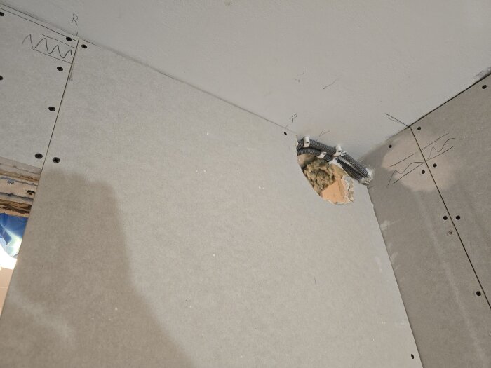 Gipsskiva med hål för ventilationsrör sitter monterad på en vägg. Rör och isolering syns i hålet, omgiven av skruvade gipsskivor och markeringar.