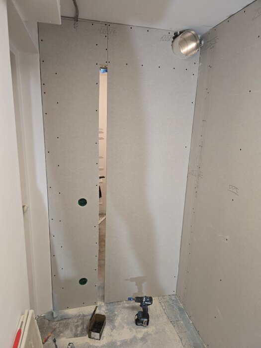 Gipsvägg under konstruktion med borrmaskin på golvet och hål för ventilation. Några skruvar och verktyg ligger också på golvet vid väggen.