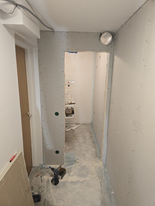 En hall under renovering med nygipsade väggar och ett ventilationsrör synligt i hörnet. Verktyg och byggmaterial ligger på golvet.