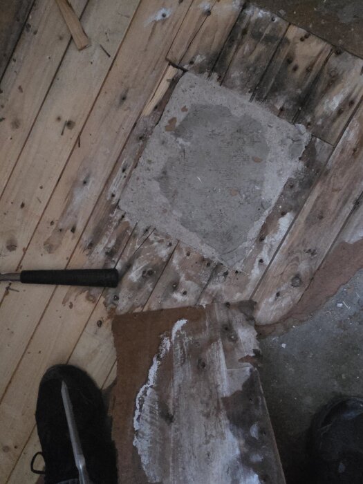 Delvis uppbrutet trägolv visar undergolv med verktyg och en sko synlig bredvid en cementplatta, del av renoveringsprojekt.