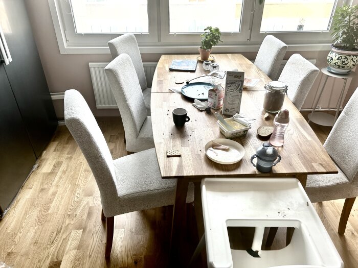 Ett matbord med stolar, tomma fat, smörpaket, mjölk, en vattenflaska och en barnstol. Servis och smulor syns på bordet med några växter vid fönstret.