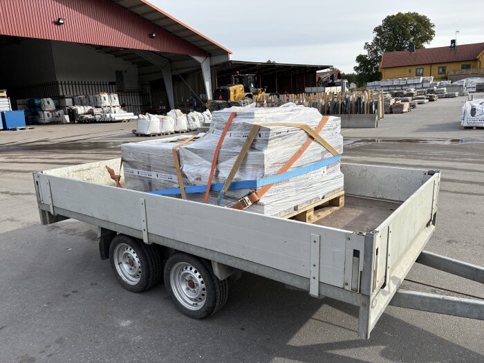 Släp med lastpall innehållande byggmaterial, säkrad med remmar, placerad på en industriell gård med lager i bakgrunden.