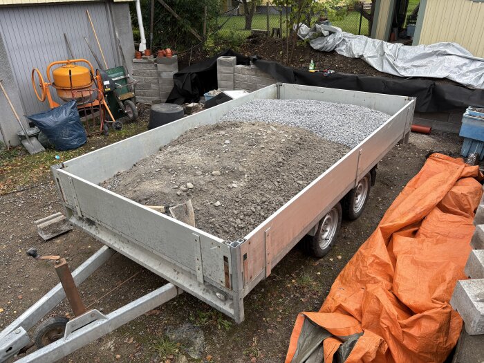 Släpvagn lastad med jord och grus står på en uppfart, omgiven av verktyg och orange presenning.