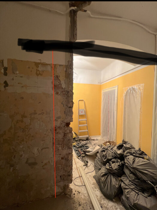 Rum under renovering med öppnad vägg för stålbalk, verktyg och svarta sopsäckar på golvet, stege lutad mot väggen i bakgrunden.