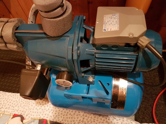 Blå vattenpump med tryckmätare och grått eluttag, isolerade rör och metallplatta på en filtad yta inomhus.