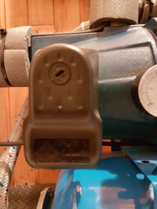 Närbild av en blå foraspump med manometer och isolerade rör i ett fritidshus, med fokus på pumpens inställningsdel.