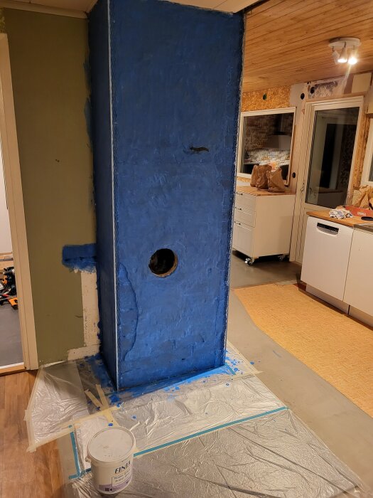 Vägg i köket med blåmålad primer och ett runt hål, omgiven av plastskydd på golvet. Primerburk med texten "Finja" synlig i förgrunden.