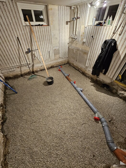 Renovering av tvättstuga med nytt avloppssystem och urgrävt golv täckt med grus. Väggen har mönstrad tapet och flera verktyg står lutade mot väggen.
