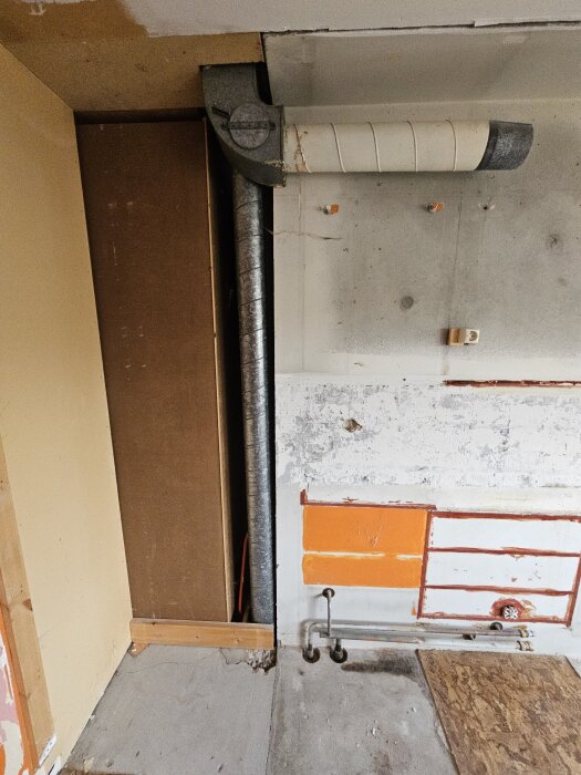 Köksrenovering pågår; synlig betongvägg med ventilationsrör, rör vid golvet och delvis orange målad yta, samt oskyddad trästrukturer till vänster.