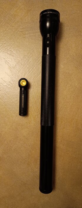 Två svarta ficklampor, en stor och en liten, ligger på ett bord. Den större är klumpig medan den mindre är en laddningsbar LED-lampa.