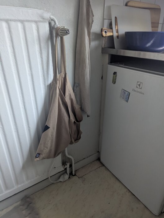 Tunn radiator bredvid kylskåp, med tygpåsar hängande, installationen är trång vid golvet.