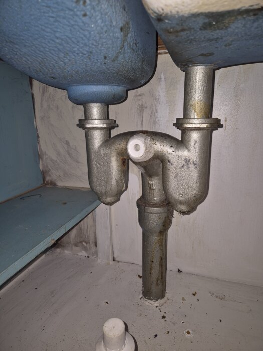 Gammalt avloppssystem i metall under diskbänk, två rör möts vid ett grenrör. Rosten syns på röret, möjligt byte till plaströr planerat.