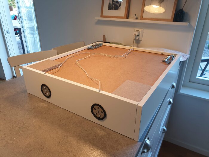 Bakre sidan av ett nedmonterat köksskåp med synliga kablar och plats för spottar, liggandes på ett köksbord i en modern inomhusmiljö.