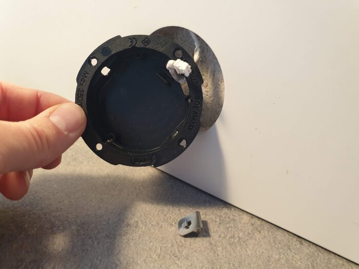 Hand håller en LED-spotlight-infästning vid en öppning i en möbelyta, liten metallklämma ligger på bordet.