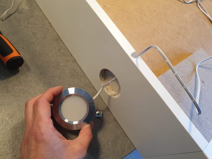 Montering av en ny LED-spot i ett skåp, en hand håller spotten med vit kabel genom ett borrat hål i skåpsstommen.