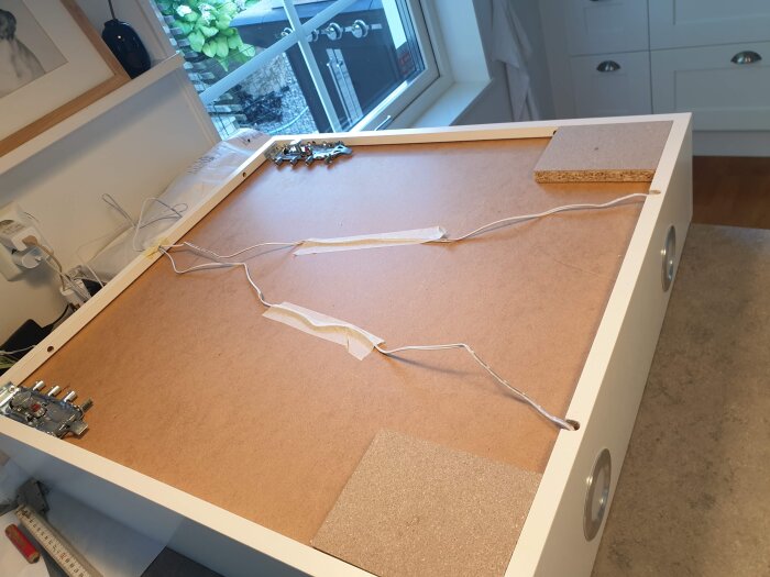 Undersidan av ett skåp, omkopplingsarbete pågår; kablar är fästa vid skåpsbotten och några delar ligger på bänken intill.