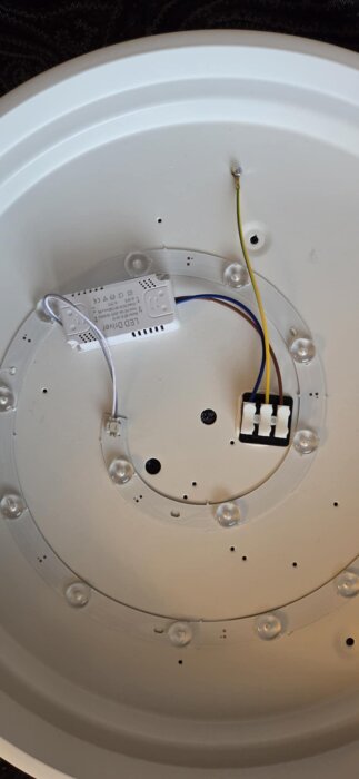 Insidan av en lampa med gul jordsladd kopplad till en skruv genom metallramen, LED-driver och kopplingslist synliga.