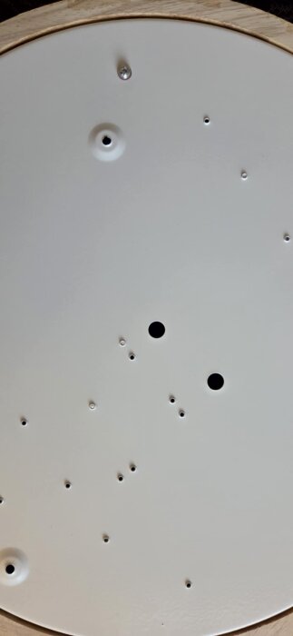 Vit lampskärm med flera små och två större hål, samt en skruv som sticker ut genom ytan.