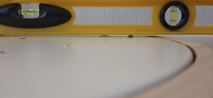 Gul vattenpass över en vit yta där en skruv sticker upp som den högsta punkten på en lampa, millimeter högre än distanserna.