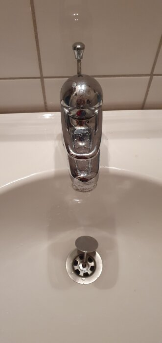 Kromat handfat med pop up-ventil och vattenkran över vitt porslin, reflekterar ljus från kakelvägg.