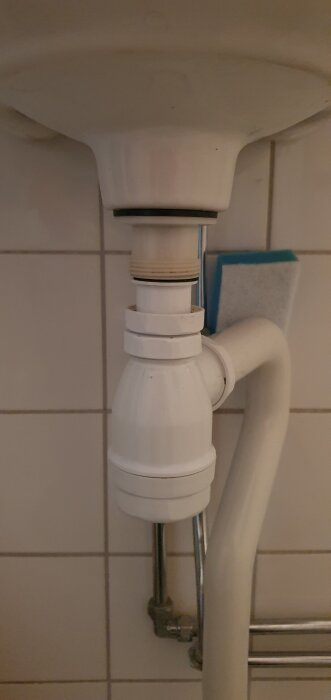 Undersidan av ett handfat med en vit avloppsventil, rör och en svamp i bakgrunden. Diskuteras byte till pop up-ventil.