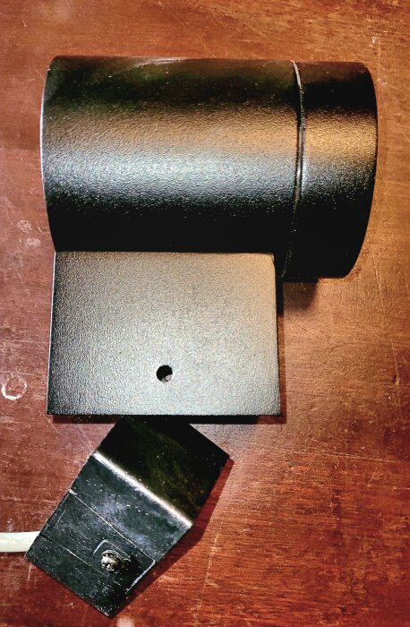 Svart spotlampa avsedd för montering på trästolpe, med fäste och hål för kablage, placerad på träyta.