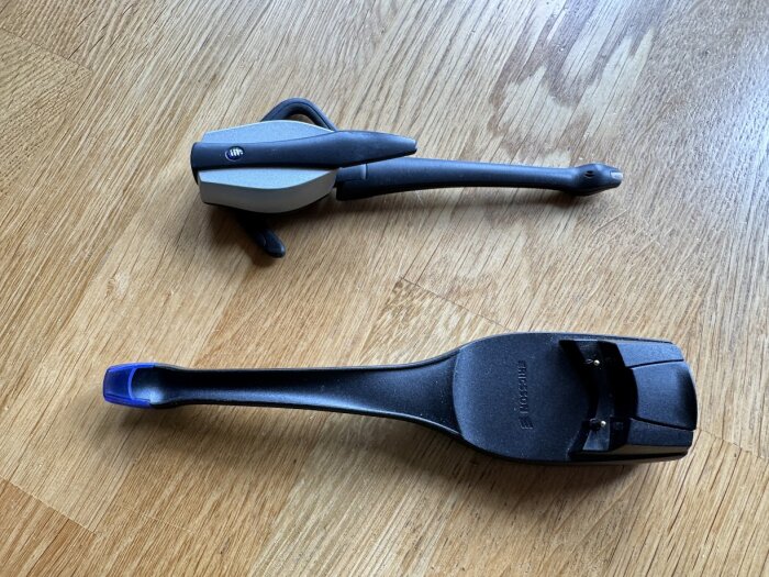 Två äldre bilhandsfree-enheter ligger på ett trägolv, den övre är silvergrå och svart medan den nedre är svart med en blå del.