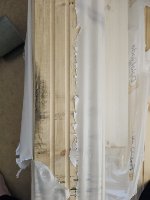 Massivt furugolv med sprickor i kvistar och delvis bortskadad ytbehandling, fotograferat med förpackningen delvis kvar.