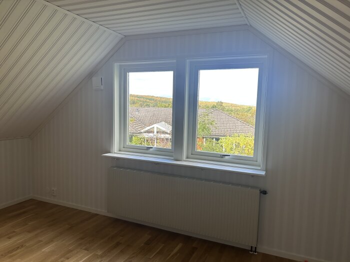 Ett allrum med snedtak och två fönster, utan gardinstänger, med trägolv och tapetserade väggar. Utsikt över ett villatak och skog.