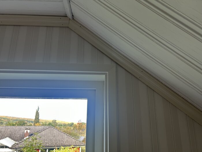 Loftfönster i ett 90-talshus med snedtak och panel, utsikt över hustak och höstlandskap.