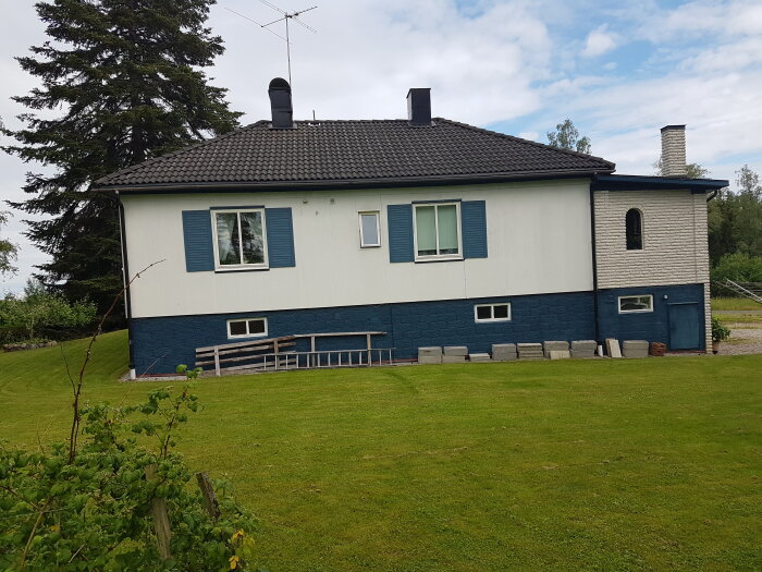 Suterränghusets baksida med gräsmatta, fönsterluckor och tegelpanel. Trädgård och träd i bakgrunden.