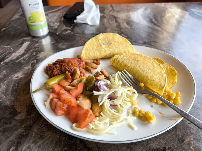 Taco tallrik med tomater, ost, paprika, majs, kyckling och sås på ett bord, burk med citrusdryck i bakgrunden.