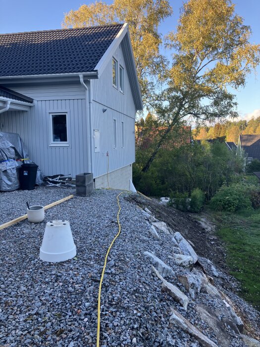 Grusad slänt vid ett nybyggt hus med en regel på marken, träd och buskar i bakgrunden. En gul sladd löper längs med slänten.
