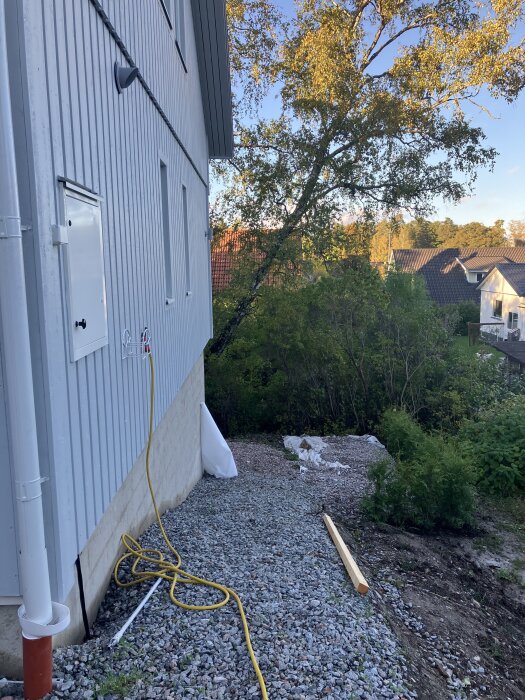 Grusad slänt vid nybyggt hus, stående regel på marken. Träd och buskar i bakgrunden samt ett gult vattenslang på marken.