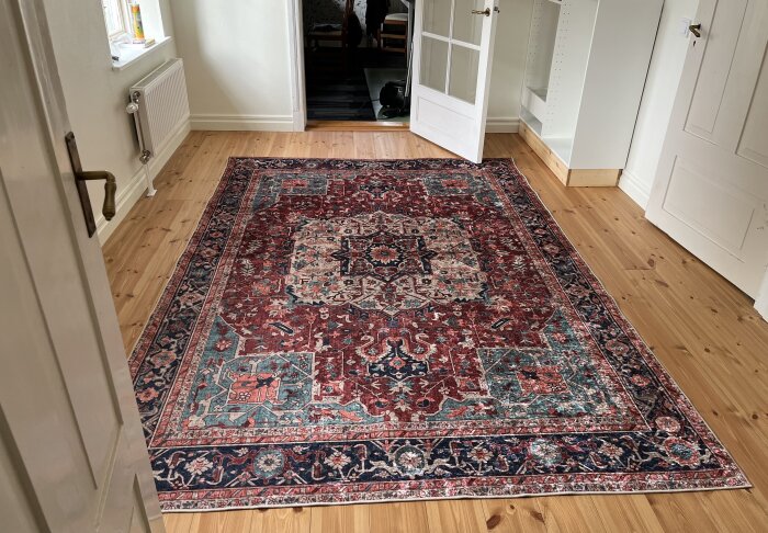 En stor, mönstrad matta i rödaktiga nyanser ligger på ett trägolv i ett ljust rum med en öppen dörr i bakgrunden.