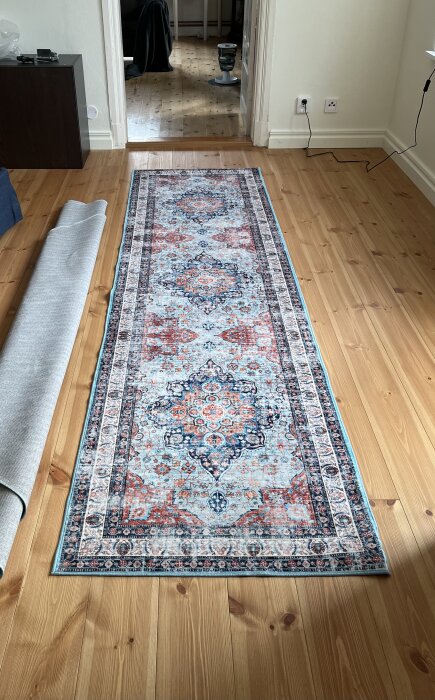 Rödaktig matta med blå och mönstrad detalj, utrullad på ett trägolv i ett rum med vitmålade väggar och eluttag i hörnet.