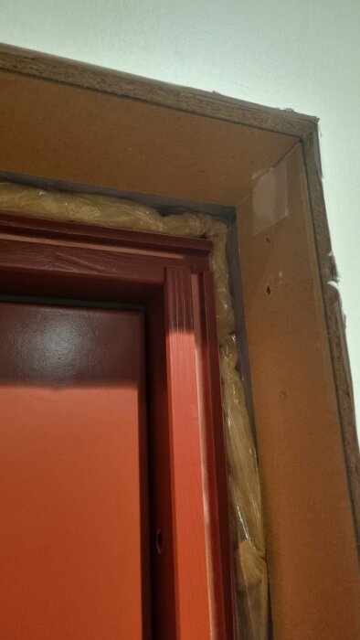 Röd dörrkarm med fräst spår, delvis monterad, omgiven av trä. Synlig isolering. Diskuterad i forumtråd om byggprojekt och dörrmontering.