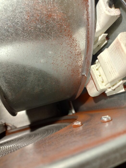 Ventilationsaggregat med rödaktigt pulver på metallkomponenter, nära anslutningar och fläkt, potentiellt tecken på rost eller annat material.