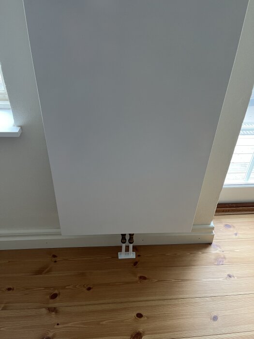En vit radiator monterad på en vägg ovanför ett trägolv, med kopparrör anslutna längst ner.