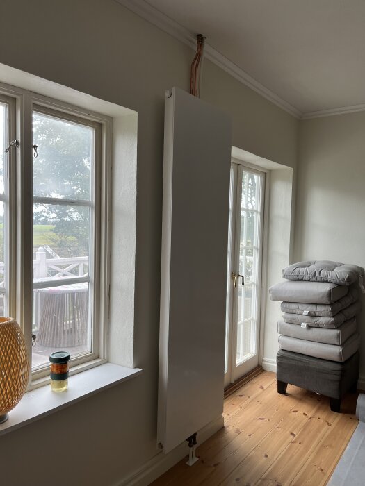 Vitt stående element monterat mellan två fönster i ett rum med träplankt golv, synliga röranslutningar ovan elementet, staplade kuddar bredvid.