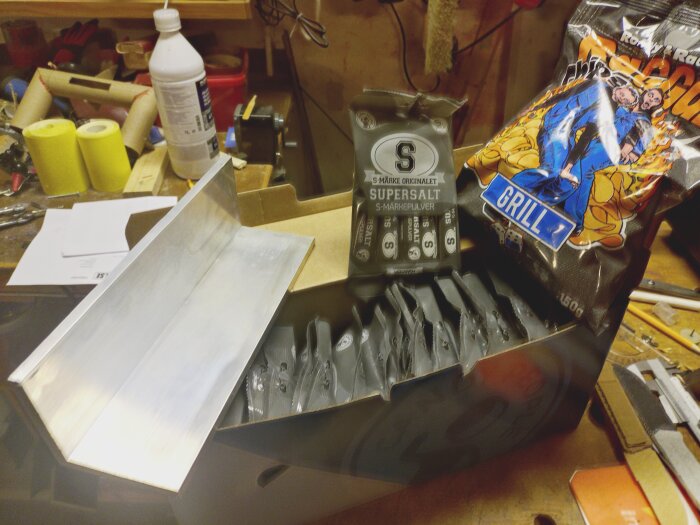Aluminiumbit på ett arbetsbord, omgiven av en kartong med supersaltpaket och en påse chips med "grill" på. Verktyg och material i bakgrunden.