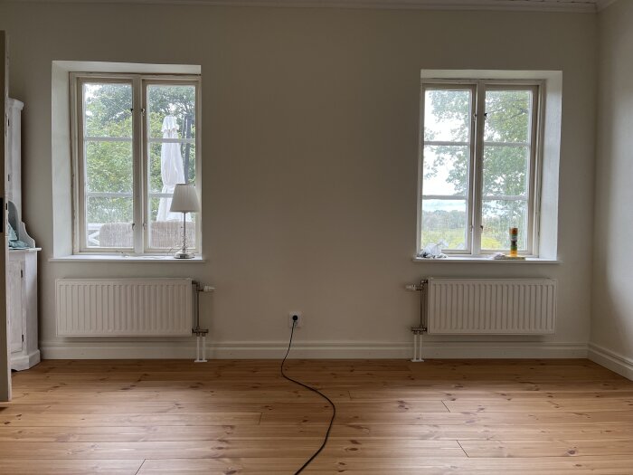 Två fönster med vita element under dem i ett rum med trägolv. Elementen och rördragningen visas i hörnen under fönstren.