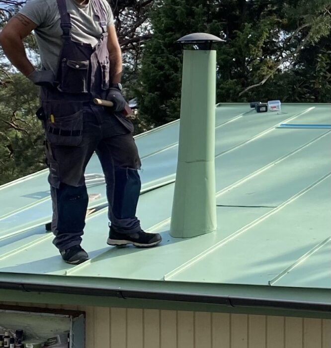 En person står på ett grönt tak bredvid en skorsten med en grön stos. Det syns verktyg på taket och omgivande träd.