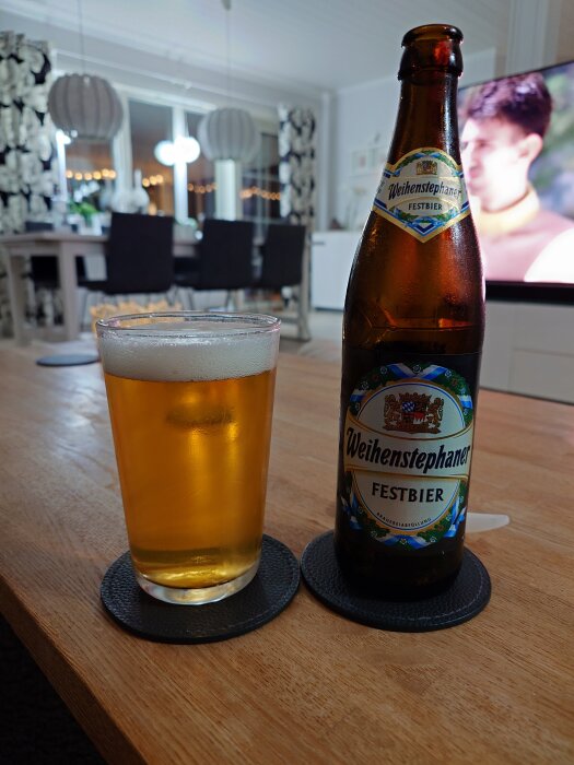 Flaska Weihenstephaner Festbier och ett glas öl på ett bord i ett vardagsrum med matplats i bakgrunden.