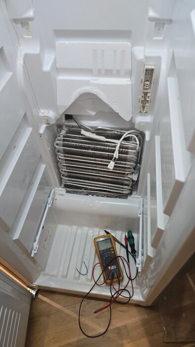 Öppnad Electrolux frys med exponerad värmeslinga, multimeter och skruvmejsel på frysgolv, ledningar anslutna för felsökning.