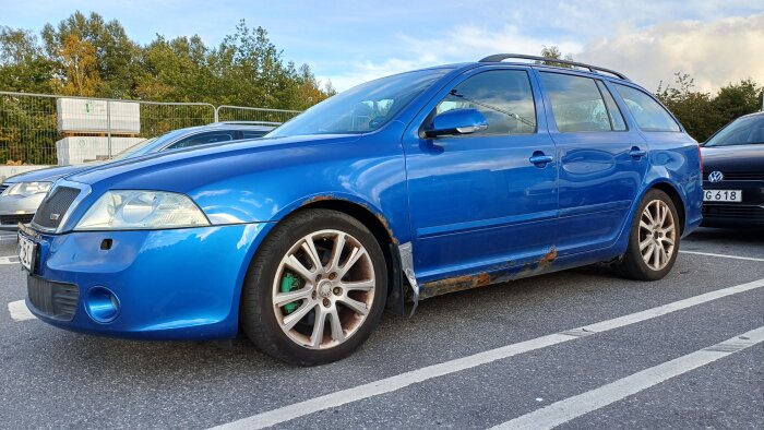 Blå Skoda Octavia från 2006 med rostskador längs hjulhus och tröskel, fotograferad på en parkeringsplats i Haninge.