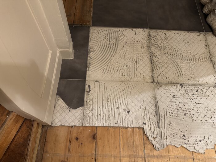 Furugolv med borttagna klinkerplattor och kvarvarande bruk, synlig träram och vägg intill.